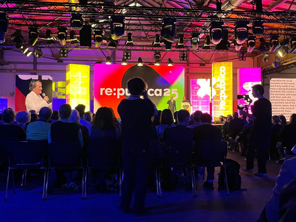 Blick auf Bühne 1, Eröffnungsveranstaltung. Unten im Bild dunkel sitzende Menschen. Die Bühne ist bunt ausgeleuchtet, Sprechende sind sehr klein oder auf einem Bildschirm zu sehen. Im Vordergrund dunkle Silhouetten von Kamerapersonen.
