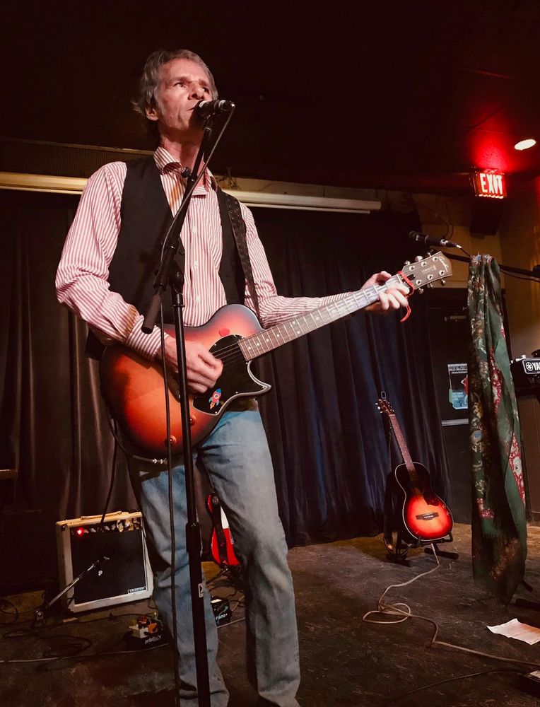 Photo of T.C. Folkpunk on stage at The Black Swan in Toronto. The backdrop is dark, with a black curtain along the rear wall, ceiling painted black, and dark grey carpet covering the stage floor. T.C. is at the microphone in mid-song, wearing a dress shirt with thin red and white vertical stripes, a black vest, and blue jeans. Behind him are two guitars on stands belonging to Maddisun and her guitarist Steve, plus a small white guitar amp belonging to Steve.