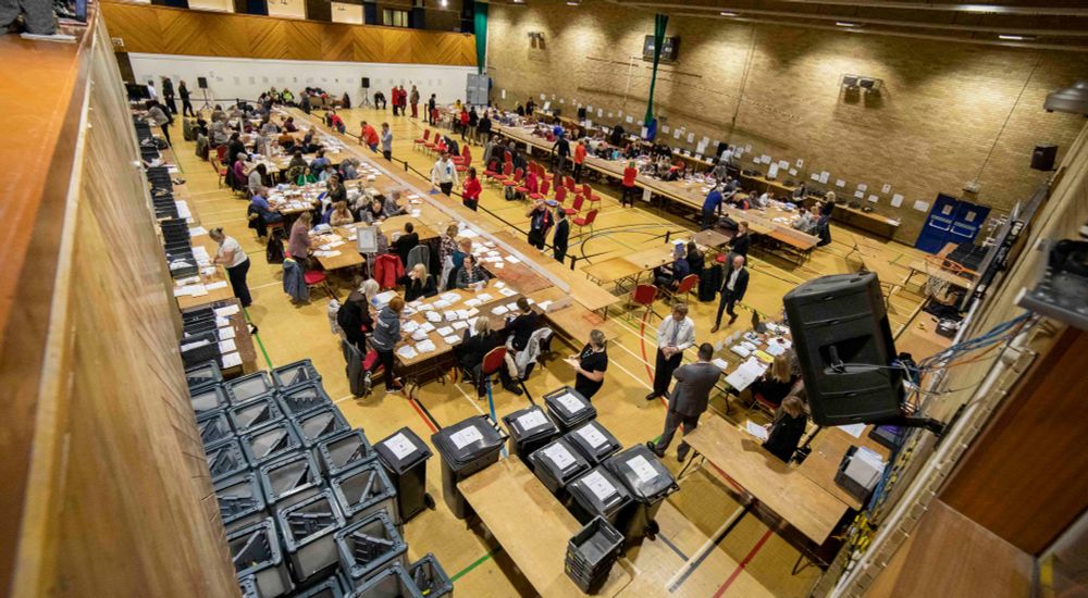 A school hall with tables and boxes around the periphery. People are casting their vote in an election. Various papers are strewn on the table. Red plastic cheers are placed in a line down the centre of the room for people to wait. 