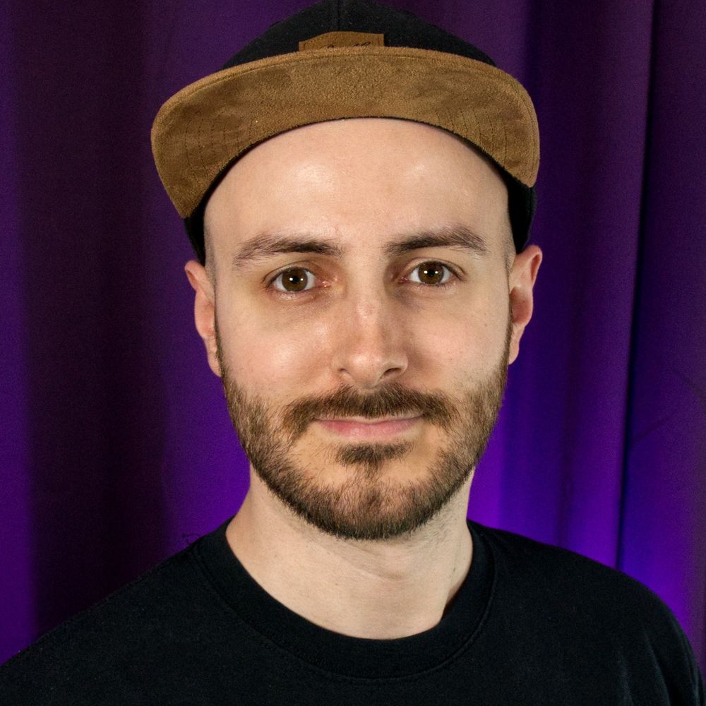 Ein Foto von Mario aka The Blake Box. Er trägt eine Kappe, Vollbart, eins schwarzes T-Shirt und lächelt mehr oder minder charmant in die Kamera. 💋