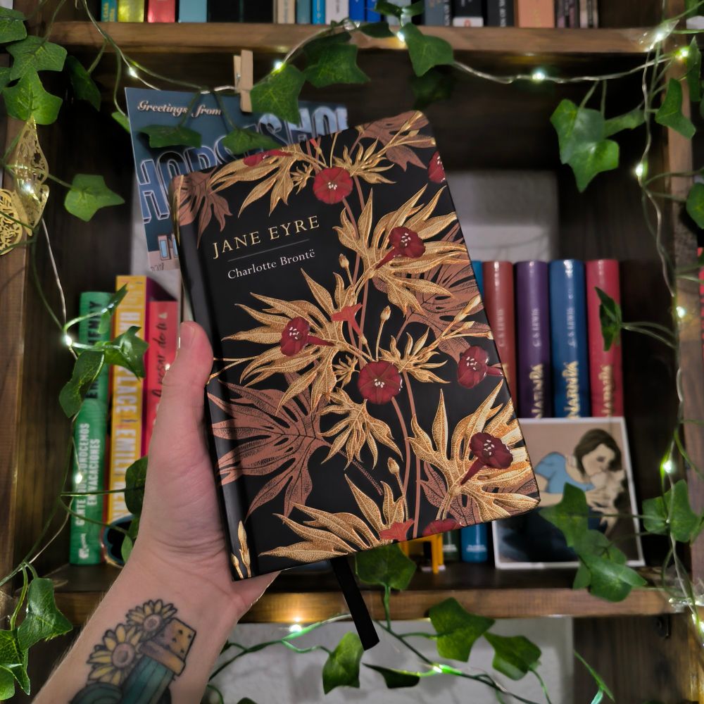 in the background there's a bookcase full of books and it has fairylights with green leaves

in the front there's a white hand holding a edition of jane eyre by Charlotte brontë. the background of the cover is black and has palm leaves and golden and red flowers on it