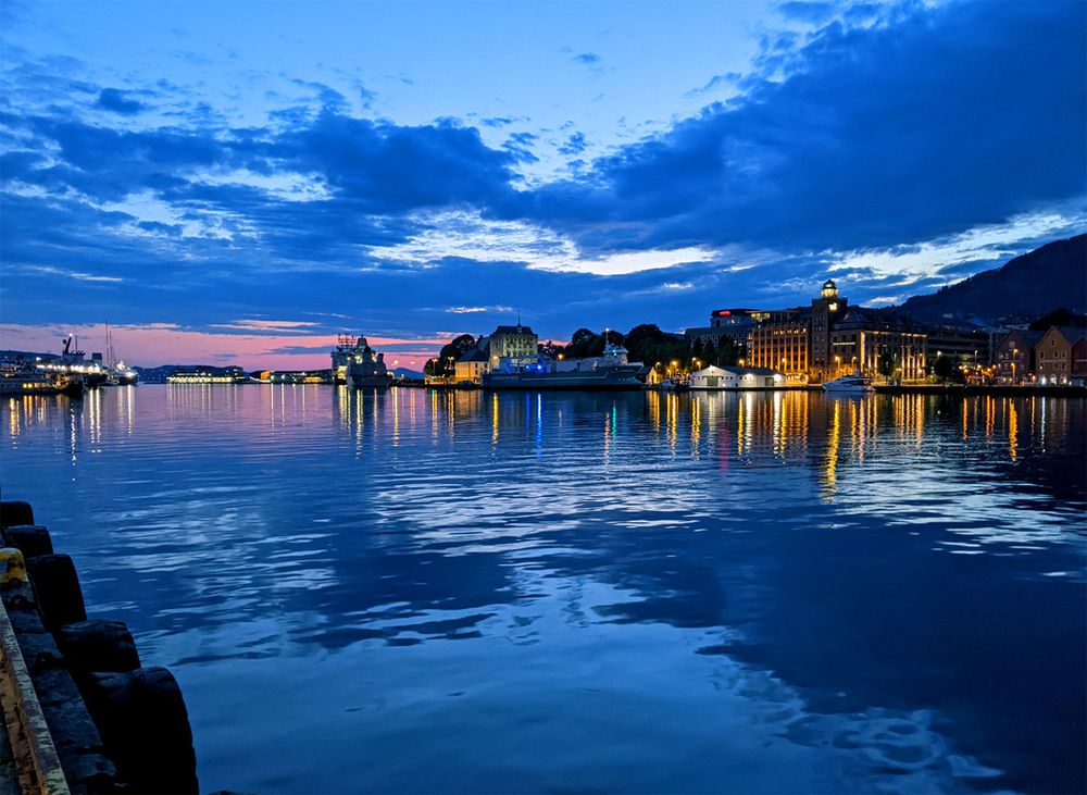 At the edge of the water under a blue twilight, with warm street and building lights reflected in the water. The sky is still pink at the horizon even though it's long past midnight.