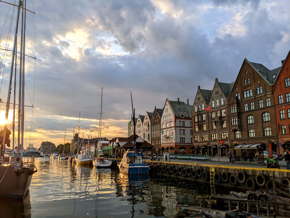 The Bergen Brygge during a cloudy golden hour, with sunlight reflecting off the water, boats, and the windows of the old buildings.