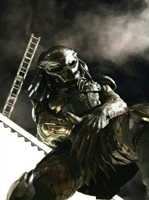 A behind the scenes picture from a Predator movie. The Yautja, with predator mask on, looks down at the camera which seems to be under its legs akimbo, the clawed hands covering at the groin area. 