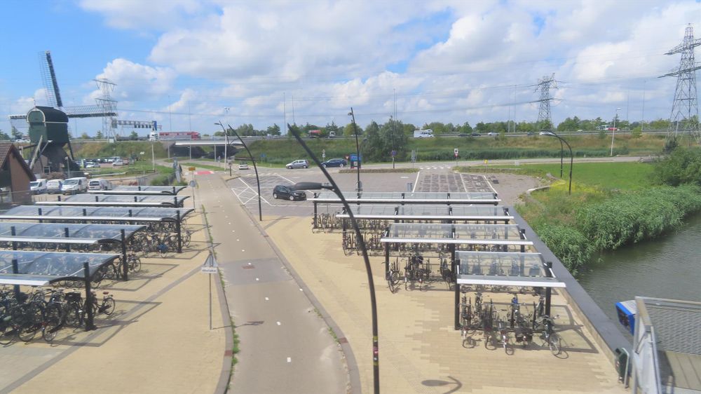 View from a double decker train as it passes over a cycleway, either side of the cycleway there is bike parking and to the left a windmill in the background 