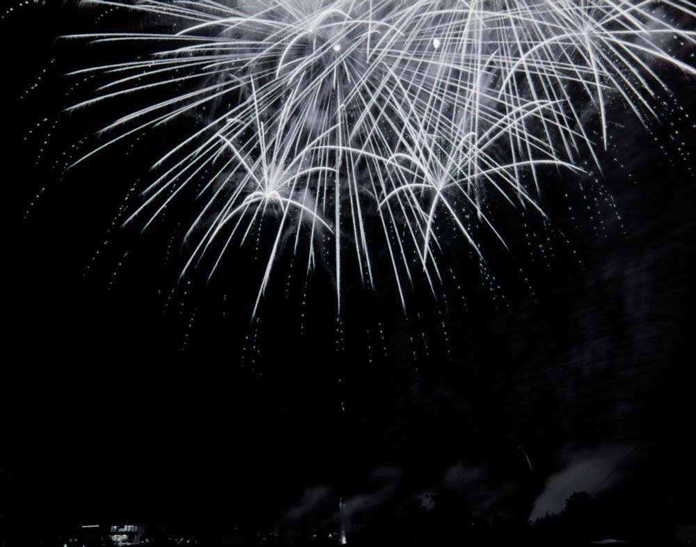 A black and white photo of 4th of July fireworks breaking overhead. Anchored at the bottom by the tops of buildings with their interior lights on and trees silhouetted by a haze of smoke. The moon can be seen through the streaks of burning stars at the top, slightly right of center. While it is black and white film, I digitally processed it in darktable as if it was color film to preserve the tone of the image stain which can be seen as the blue-green tone on the edges of the stars as they burn out.