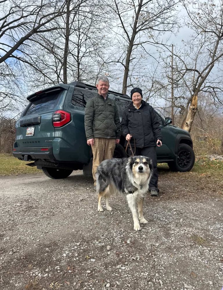 A man and a woman stand in front of a green Toyota 4-Runner and behind a black, tan and white dog. The dog is smiling at the cameraman.