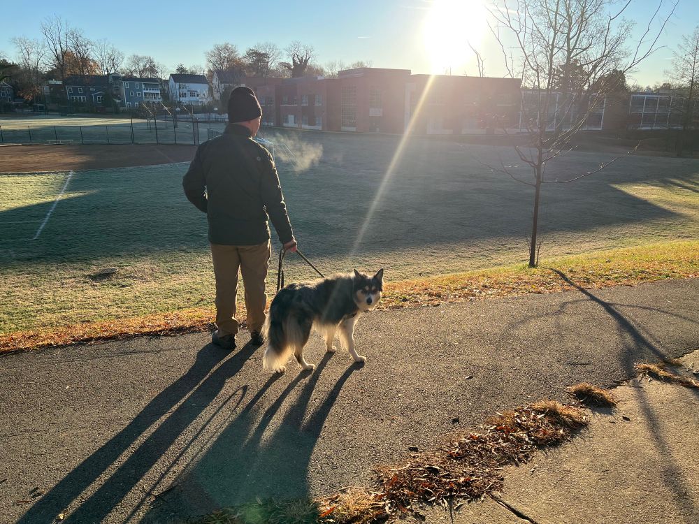 The divine light of the sun points towards a black and white dog held on a leash by a man as they stand on a hill overlooking a large field and building in the background. So cold you can see the breath of the man.