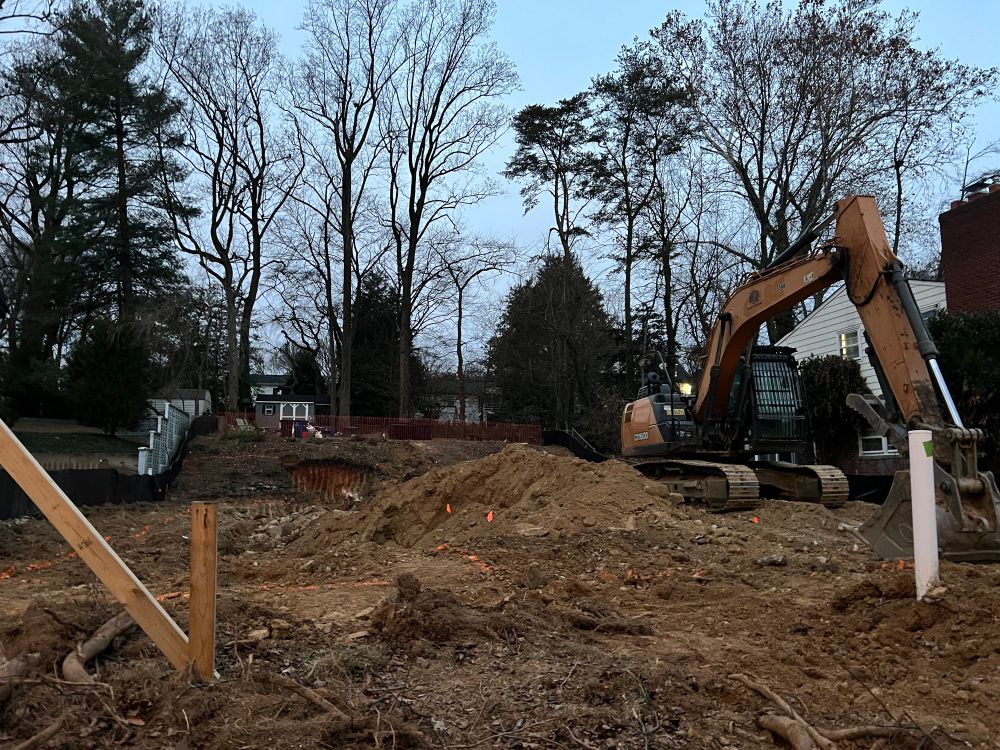 Large yellow excavator sits at rest beside a large hole in the ground where a house used to stand.