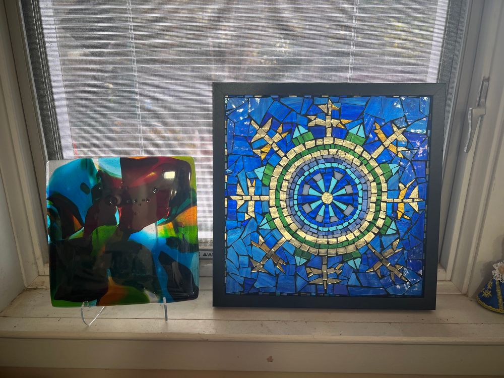 Two pieces of glass art sitting in a window. On the left is a flow fusion piece made of red, green and blue transparent glass and on the right is a stained glass mosaic of circles and spike proteins in gold, blue and green glass.