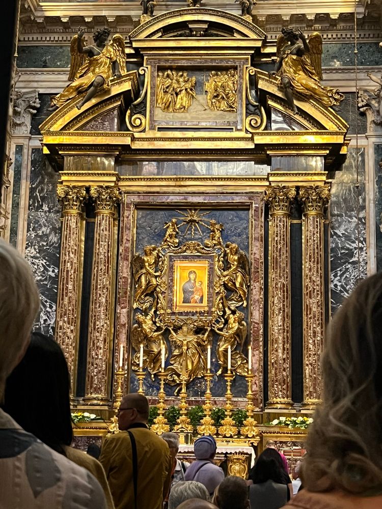An ornate mosaic of a woman and child surrounded by gold metalwork is placed between two decorated columns on the left and two columns on the right. Five candles on candlesticks are placed below the mosaic. Heads of people are in the front. 