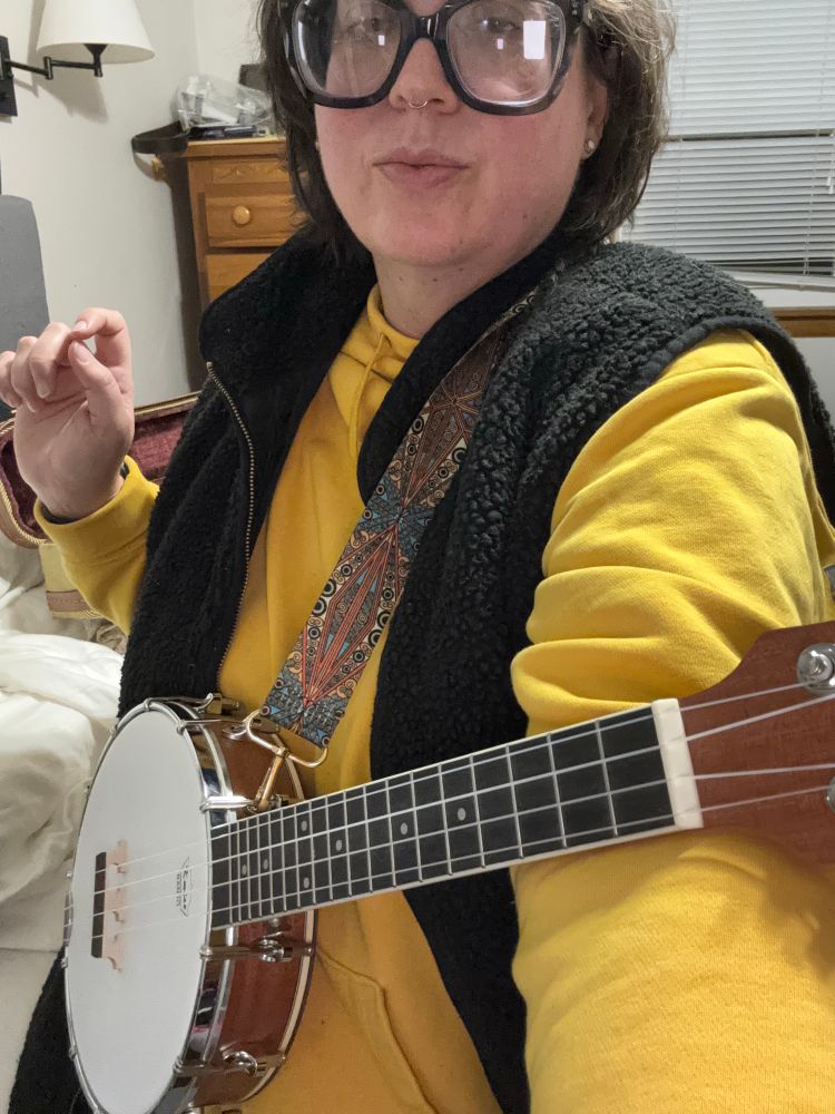 Kmise 4 string banjo ukulele, worn by me in yellow sweatshirt and black fuzzy vest. In a bedroom. 
