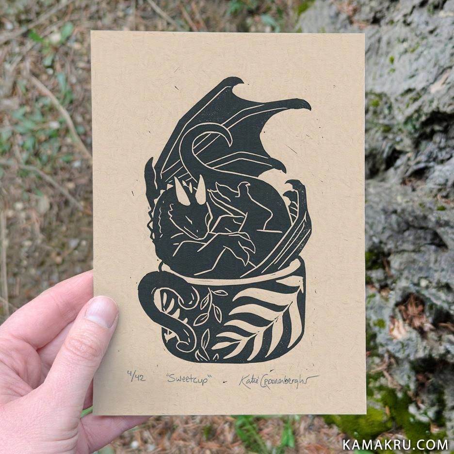 A 5x7 inch lino print in black ink on toned tan paper of a small cute dragon sitting in a wide mug. On the mug are images of ferns and leaves. Limited edition, written under the print says 4/42 the title "sweetcup" and artist signature Katie Croonenberghs. Text in lower right of image reads kamakru.com