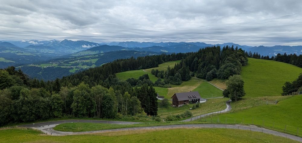 Panoramic shot of some mountains with green grass and trees. In the foreground a road meanders downwards past a house. The mountains turn more blueish towards the horizon. The sky is clouded.