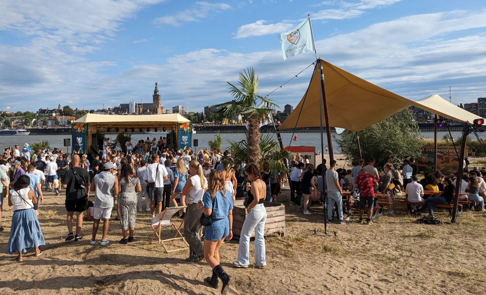 A beach with a stage on the left in front of the river. Above the canopy you can see St. Stevens church at the other side of the river. On the right is a terrace with a canopy made of cloth. In the middle a palm tree. People are walking, salsa dancing and drinking (probably beer from the local brewery Oersoep). Festival 't Eiland.