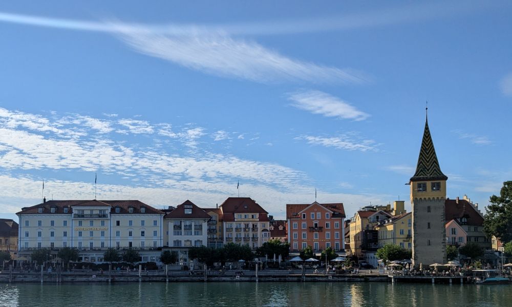 Skyline of the city of Lindau at the Bodensee in Germany with some ancient, colored buildings at the other side of the water of the harbour and the yellow Mangturm in the evening sun. The sky above is clear blue with some wispy clouds.