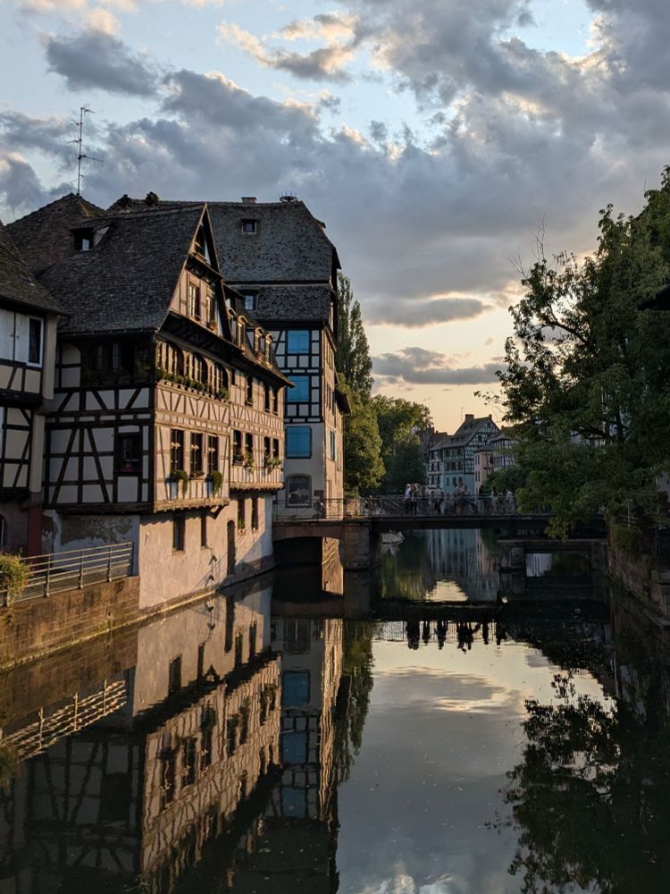 The evening sun shines on some ancient houses on the left next to the canal that runs through the city of Strasbourg. Some people stand on a bridge above it, their reflections to be seen in the still water. A large tree on the right. The sky is clouded. Houses reflected in the still water too.