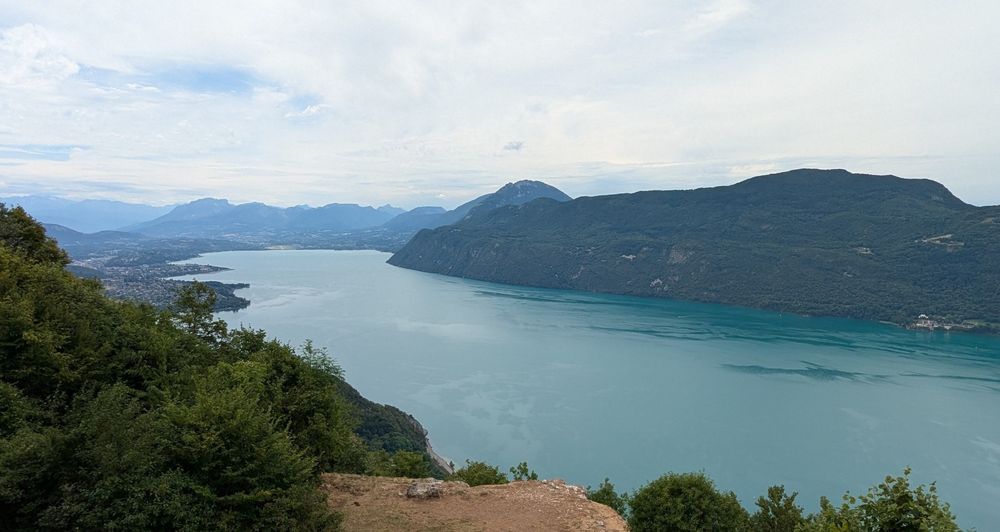 A turquoise lake stretching to the left to the horizon. Green hills at all sides and a sand colored stroke in the foreground. The sky is white from clouds.