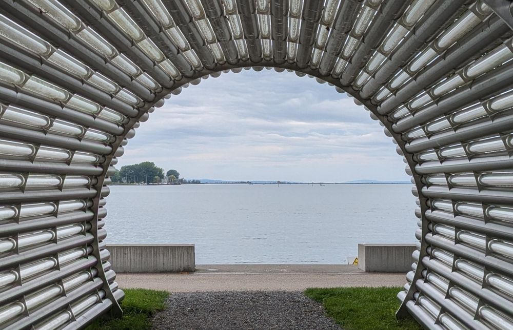 Peek through an installation made of tubes put together making a big tube you can walk and see through and look at the large lake; Bodensee. There's grass on both sides of the path through it. Concrete benchee behind on the boulevard. There are some trees in the background. The sky is clouded.