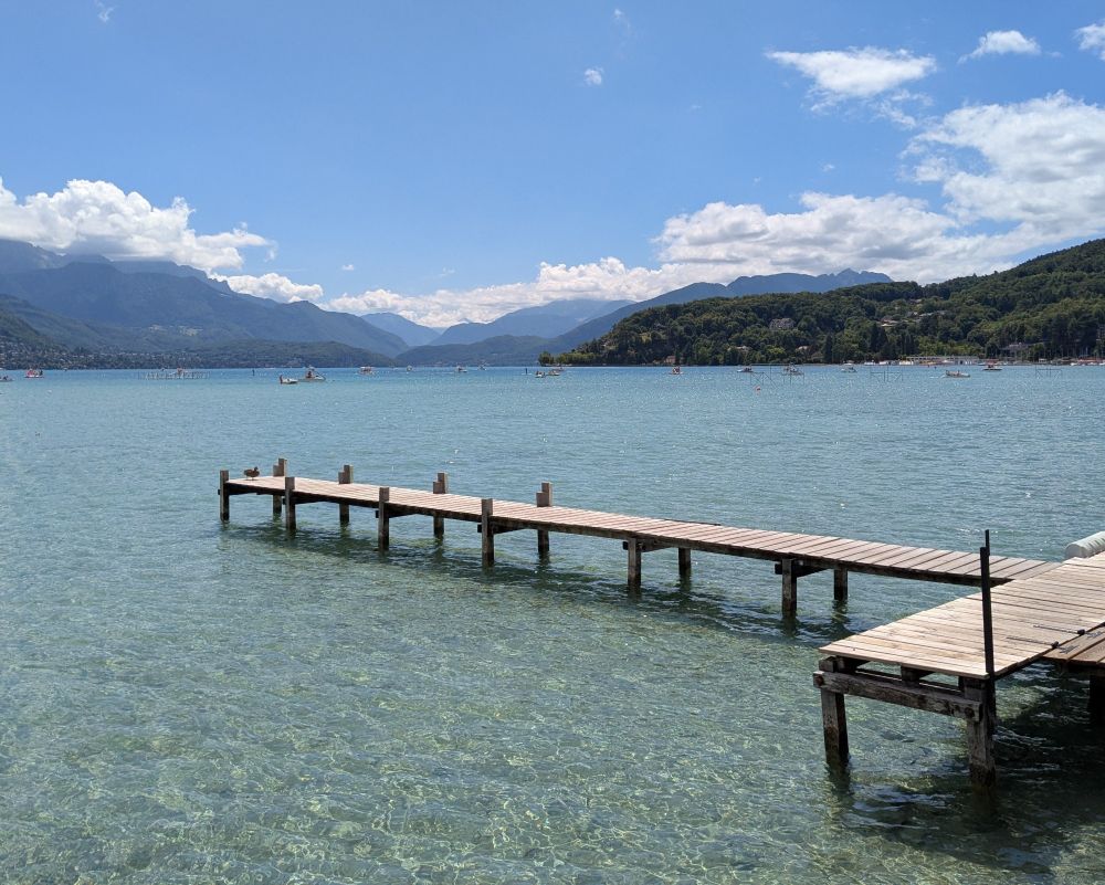 A wooden dock leading into the crystal clear waters of Lac d'Annecy coming from the right. There's a duck at the end. Layered hills in the background filling the horizon with some clouds above, below a clear blue sky. Some boats in the background as well.