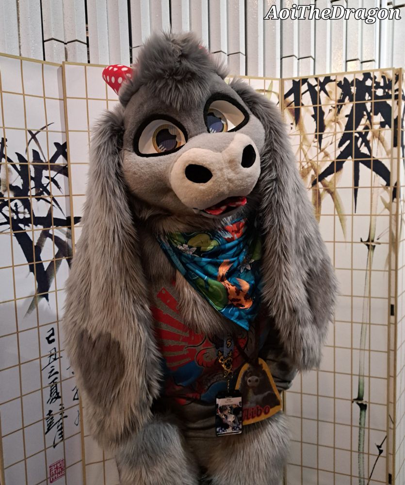 Photo of a gray plush dragon fursuiter at NordicFuzzCon 2025 inside one of the convention hotels while standing in front of a Japanese themed backdrop with bamboo on it.