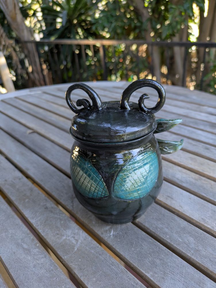 photograph of a sculpted jar sitting on a wooden table. the jar is carved an sculpted to look like the stylized face of a dragonfly with large eyes on the body of the jar, curly antennae as the "knob" on the lid and 2 pairs of wings on the back.

the jar is a very dark teal and the eyes/wings are a gradient of light teal to turquoise .

this view is showing the front
