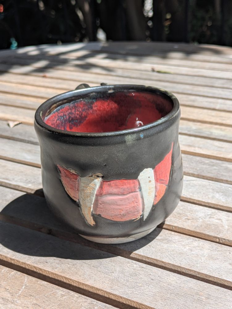 photograph of a handmade black and red ceramic mug carved with a big grin that exposes its two upper canine fangs (like a vampire) and red tongue.
the body of the mug is matte black and the inside is a gloss red. bat wings with red membranes sprout out the back of the mug and arc over the handle.

front view
