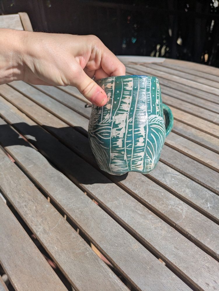 photograph of a ceramic mug with green underglaze that has had a deer like unicorn esque creature carved into it (sgraffito) amid dogwood trees.