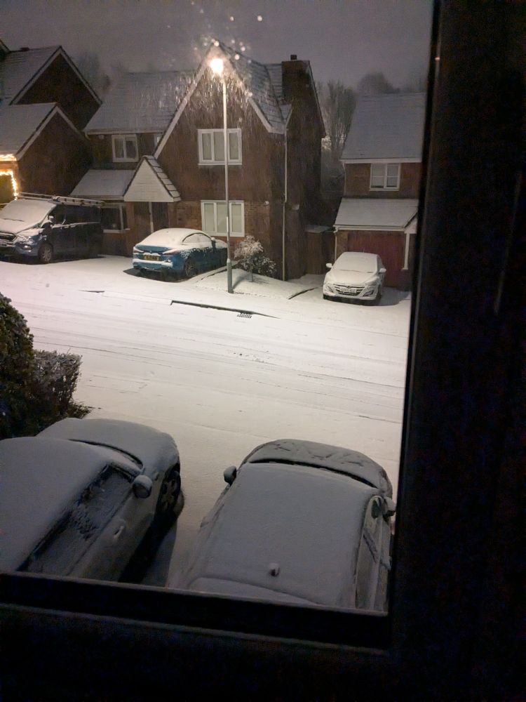 A photo of a residential street with snow covering the road, pavement and car roofs. Snowflakes can be seen around the lamppost light. 