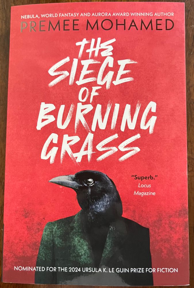 A paperback book, The Siege of Burning Grass, by Premee Mohamed. Theu cover shows a bird-headed humanoid figure in a green coat against a red background.