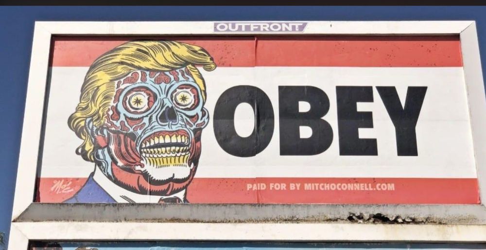 Billboard with massive lettering of the word OBEY beside caricature of face with no skin, and hair resembling FOTUS’ toupee. Has an allusion to a popular figure from a film.  