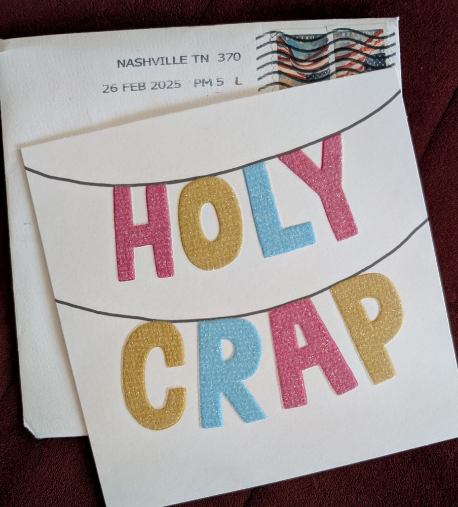 sitting on top of a postmarked and opened envelope is a greeting card with a banner made of sparklyletters in pink, gold, and sky blue that spells out HOLY CRAP. 