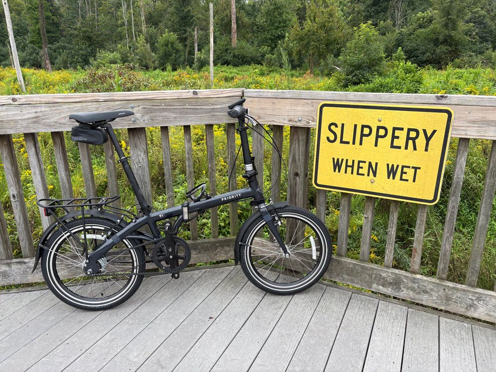 A picture of a folding bike resting against a wood railing, next to a big yellow sign saying “SLIPPERY WHEN WET”. In the background is a lush marsh with lots of little yellow flowers. 