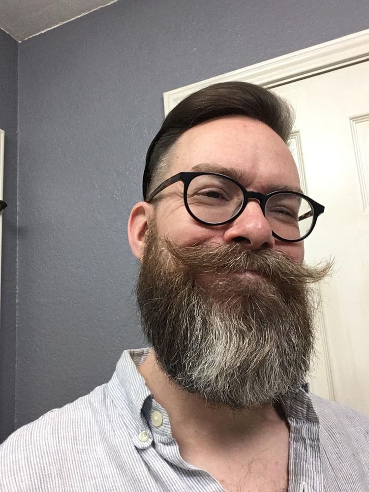 a pretransition woman wearing glasses and a blue & white striped oxford shirt. Light brown hair faded on the sides, a handlebar mustach and big greying beard