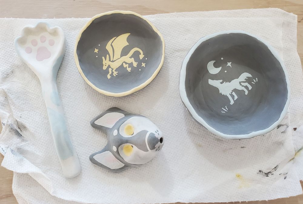 a ceramic spoon in the shape of a cats paw, white with blue accents
two ceramic bowls glazed black, one with a yellow dragon in the center of the bowl, and the other with a blue wolf 
and a howling wolf head with a mouth hole for holding incense 