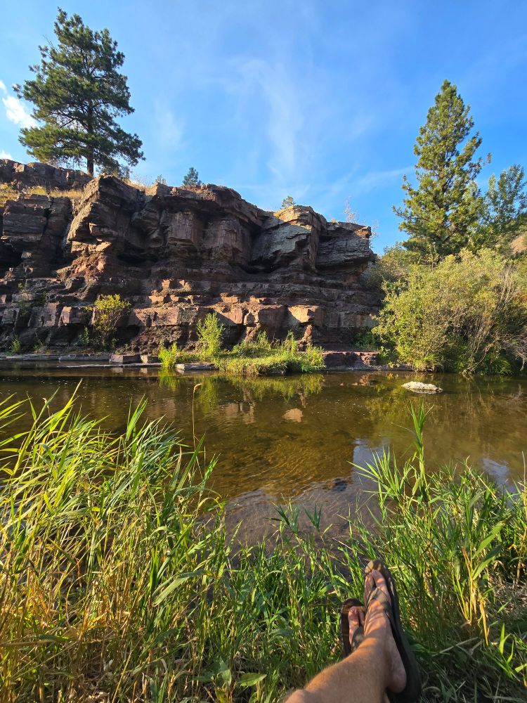 A creek passes by a rock cliff with a few tall pine trees atop. My feet can be seen in the picture as I am sitting along the bank after a cool swim.