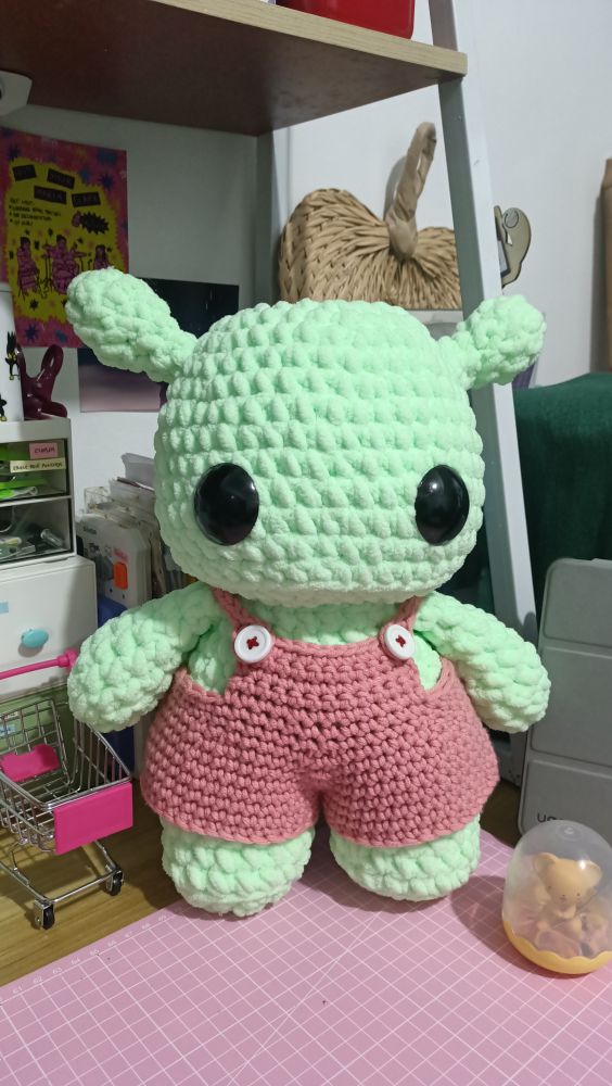 amigurumi crochet plushie of a chubby mint green alien wearing salmon pink overalls