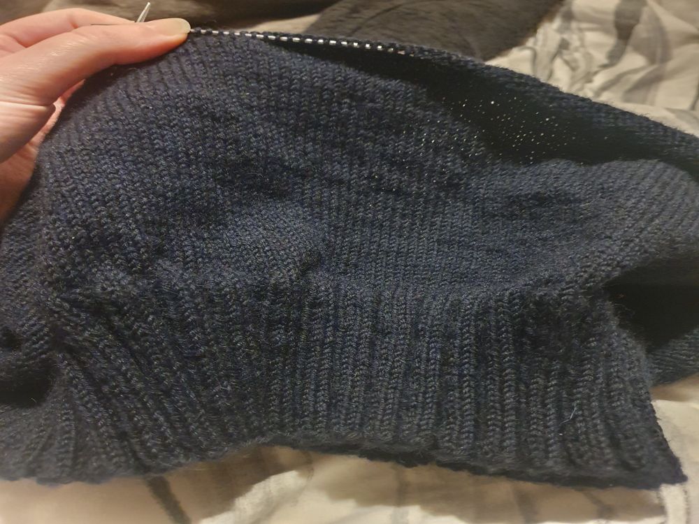 Part of a dark navy blue jumper on some knitting needles. Sits against a grey and white swirling bedsheet background