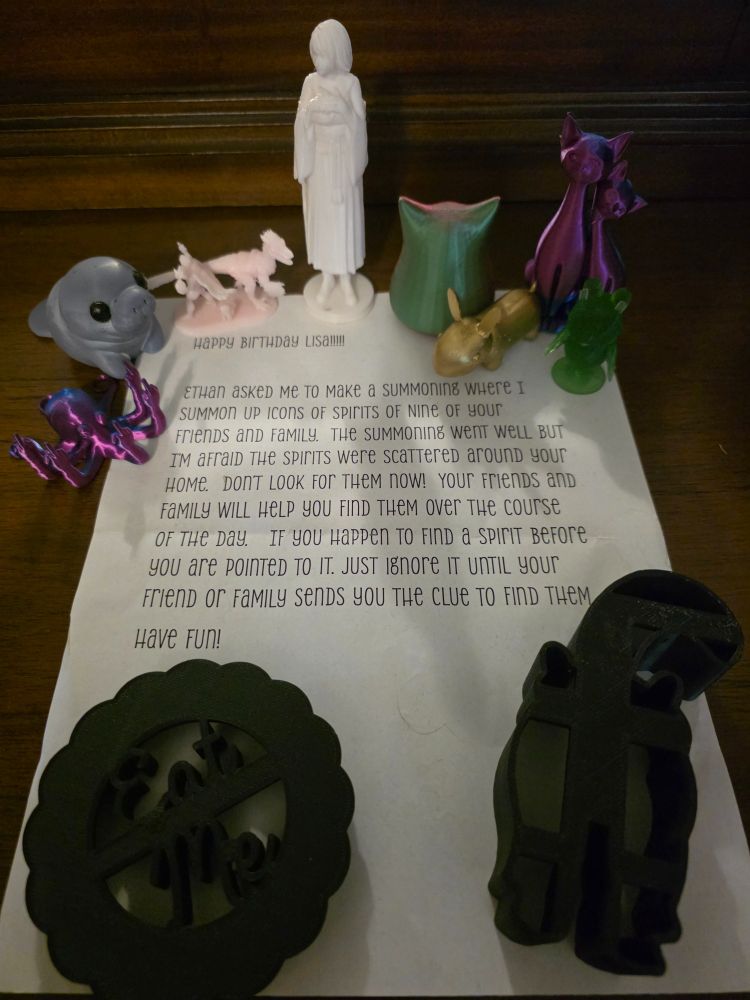 A paper that says "Happy birthday Lisa!!!! Ethan asked me to make a summoning where I summon up icons of spirits of nine of your friends and family.  The summoning went well but I'm afraid the spirits were scattered around your home.  Don't look for them now! Your friends and family will help you find them over the course of the day.  If you happen to find a spirit before you are pointed to it, just ignore it until your friend or family sends you the clue to find them. Have fun!" The figures starting at top left are a purple octopus giving the finger eight times, a grey manatee, a figurine of Dungeon Crawler Carl, mongo, and Princess Donut, Yuna of FFX, a representation of a cat, a golden nug from dragon age, a pair of purple cats, a green owl who is Celeste from Animal Crossing, and two cookie cutters: one says eat me and one is a cat butt.