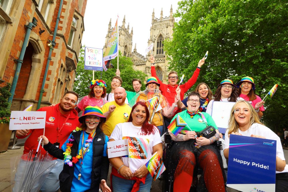 A group of colleagues from LNER, Northern and Transpennine in front of York Minister celebrating Pride with flags which read LNER Living Proud Together and Hapoy Pride