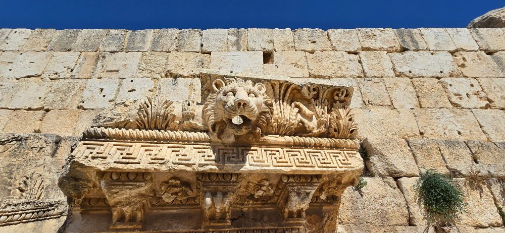An ancient stone wall is the backdrop to a huge piece of masonry with intricate carvings and a large lion's head with a hole in the mouth that was used to drain water from a Roman temple roof.