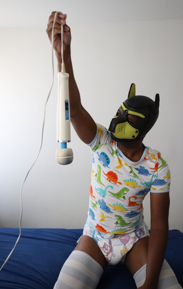 I've climbed on the bed to get a closer look! I'm pinching the wand's wire, holding it up while I examine it with a confused look on my face. I've got blue-and-white stripped socks on, and it's also revealed that my onesie is unsnapped, showing that I'm wearing a TinyTails diaper.