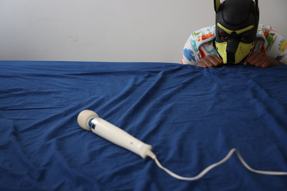 I'm wearing a yellow pup hood and a colorful dinosaur one while peering over the edge of the bed to examine a Hitachi magic wand placed on it.