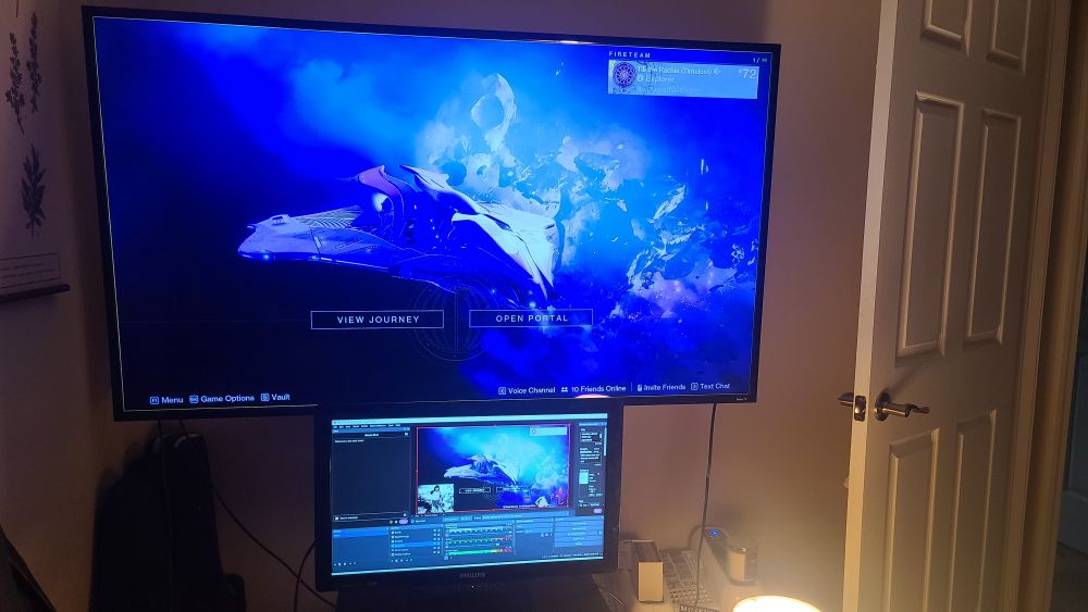 a dimly lit picture showing a huge tv screen with destiny 2 loaded up, mounted on a display stand. a tv tray holds up a second monitor, much smaller, showing an obs set up