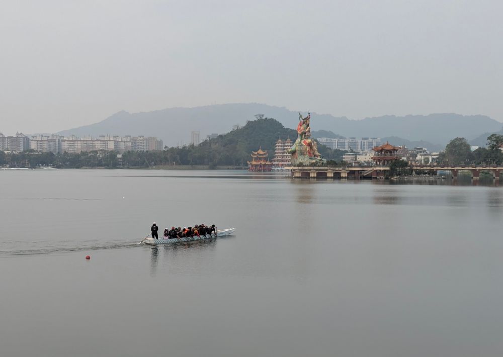Dragon boat going across a lake with a temple in the background, early in the morning.