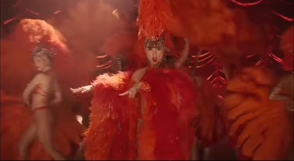 Taylor Swift in a scene from "life of a showgirl" leading single The Fate of Ophelia. This comes right after the scene where she's lying down covered and ropes and two men are putting make-up and fake eyelashes on her so she could become a showgirl again. On this scene, Taylor is dressed like a proper showgirl with lots of make-up and an orange bedazzled bodysuit and hairpiece and sleeves and hair covered and feathery very bedazzled and glorious like.
