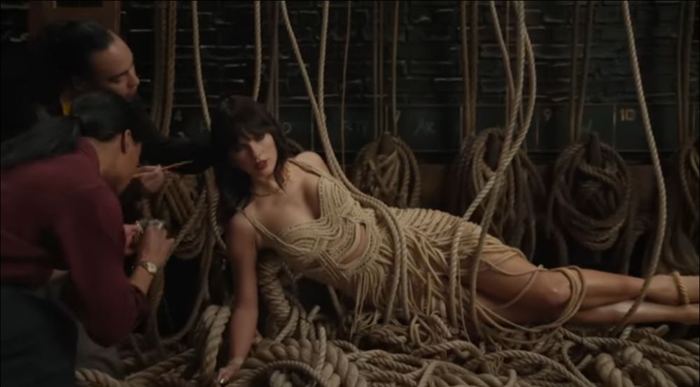 Lying down Pinocchio mode Taylor Swift on her new music video The Fate of Ophelia from her new album The Life of a Showgirl. Taylor with a black bobcat wig covered in ropes and two men are putting make-up on her and fake eyelashes. She seems depressed.