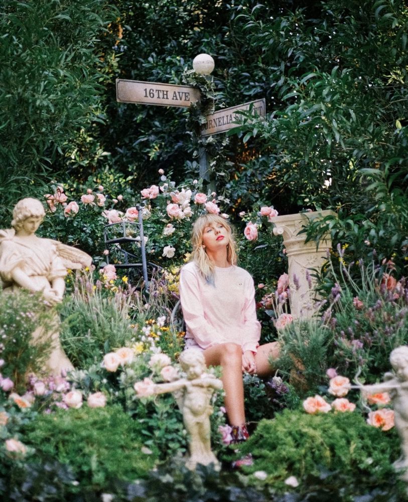 Taylor Swift sitting in a garden full of flowers from the lover album photo shoot. It's a pink and orange roses too many to count and it very much. Resembles the photos from the engagement announcement of Taylor Swift to Travis Kelce.