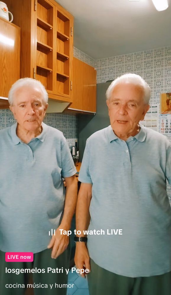 A screenshot from a live TikTok of an account of two elderly Spanish brothers (twins), wearing the same clothes, a light, blue shirt and green long trousers they are standing in their kitchen and talking about cooking in music and make jokes