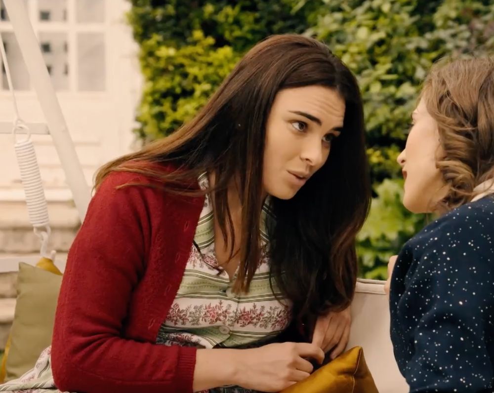 Fina in their garden talking to Marta. She is wearing a striped green, white and red dress and a red wool sweater. 